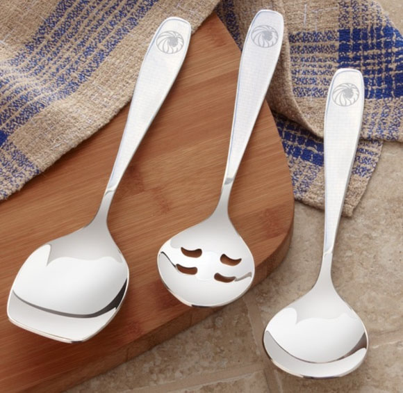 Three stainless steel kitchen utensils with decorative holes cut out to resemble smiling faces, resting on a wooden cutting board beside a striped towel.